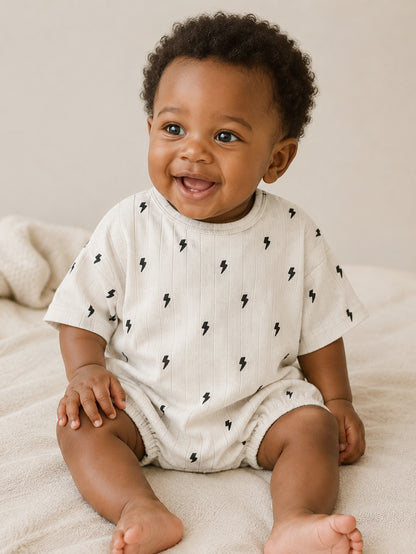 Smiling baby in a light outfit with lightning bolt patterns, sitting on a soft surface.