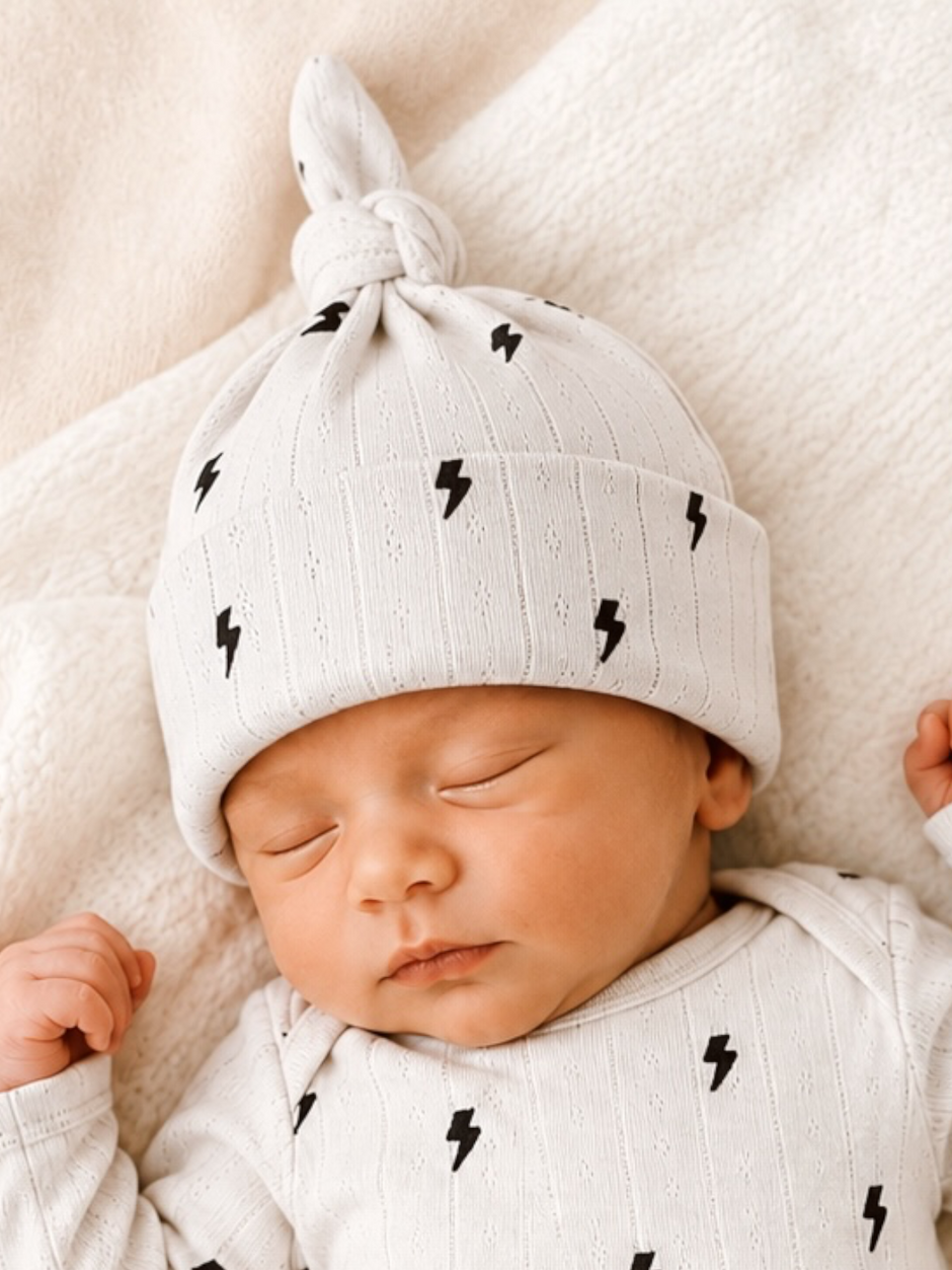 Sleeping baby wearing a light gray hat with black lightning bolt patterns, resting on a soft blanket.