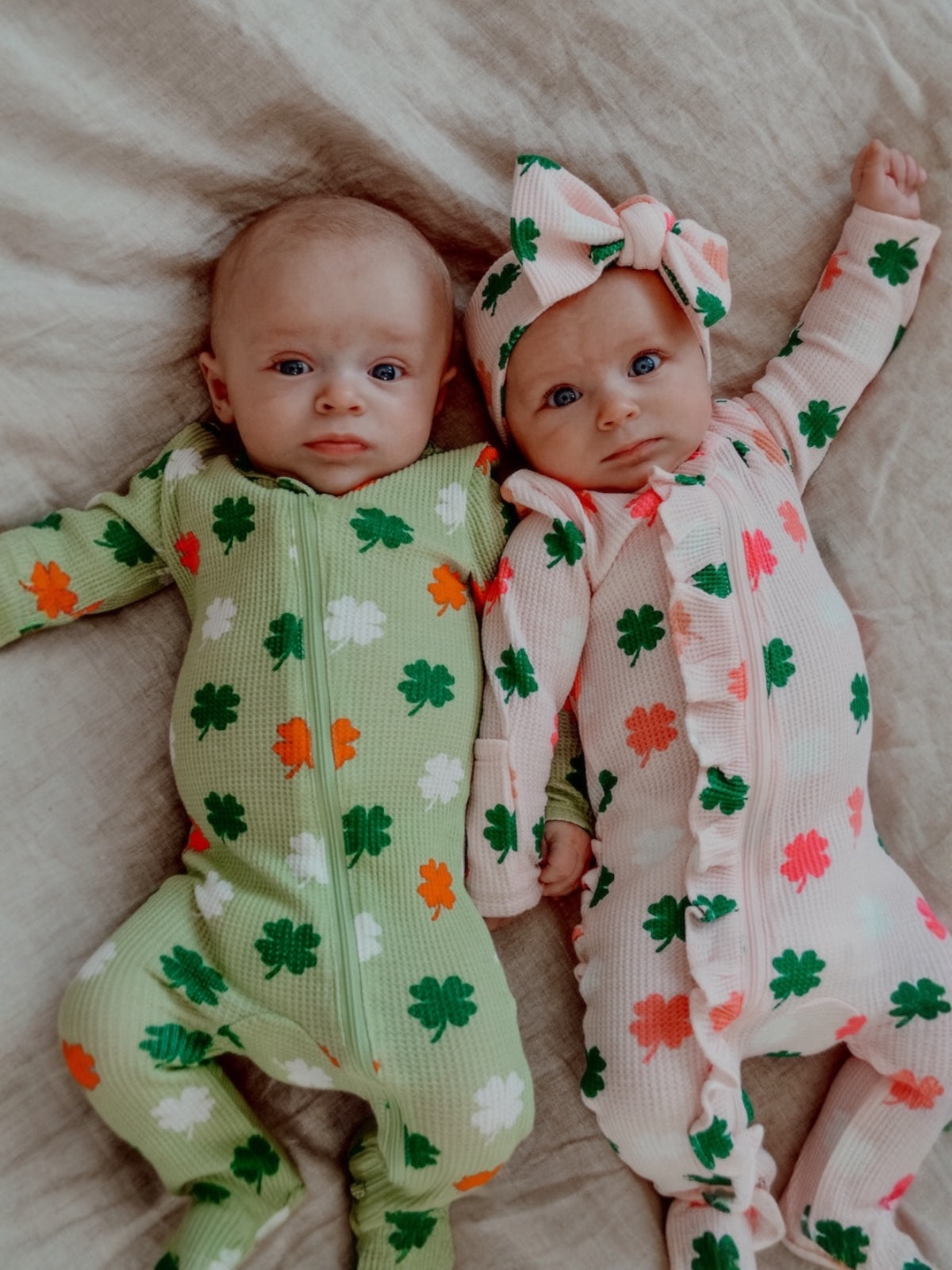 Two babies in colorful shamrock-patterned onesies, lying side by side on a beige blanket.