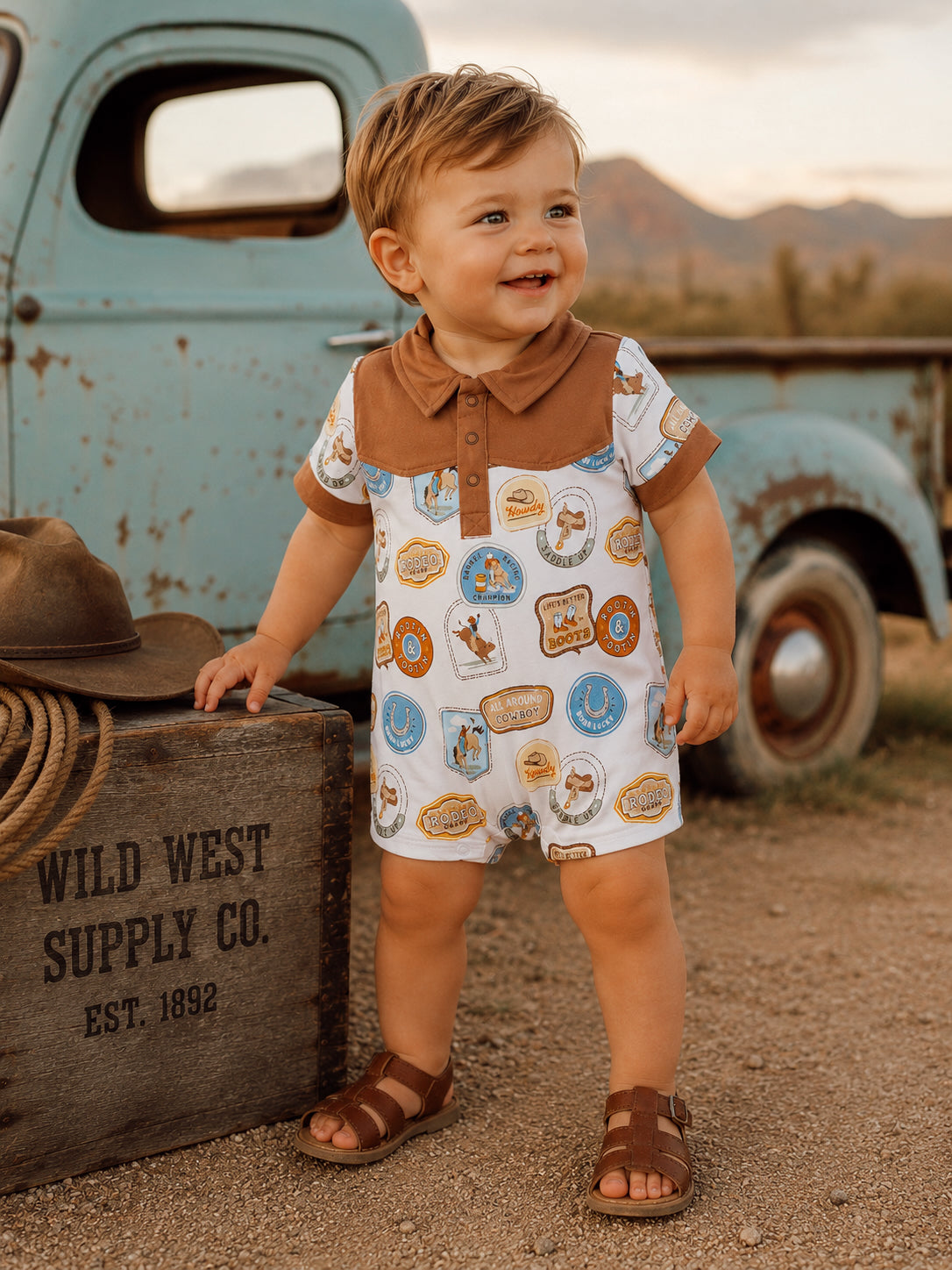 Smiling toddler in patterned onesie stands by a vintage truck, surrounded by rustic scenery and a wooden crate.