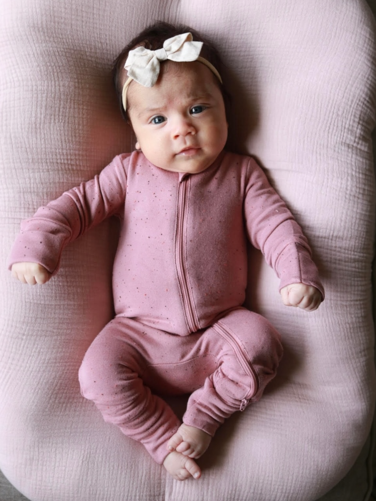 Infant wearing a pink zip-up outfit and bow headband, lying on a soft pink surface, looking curiously at the camera.
