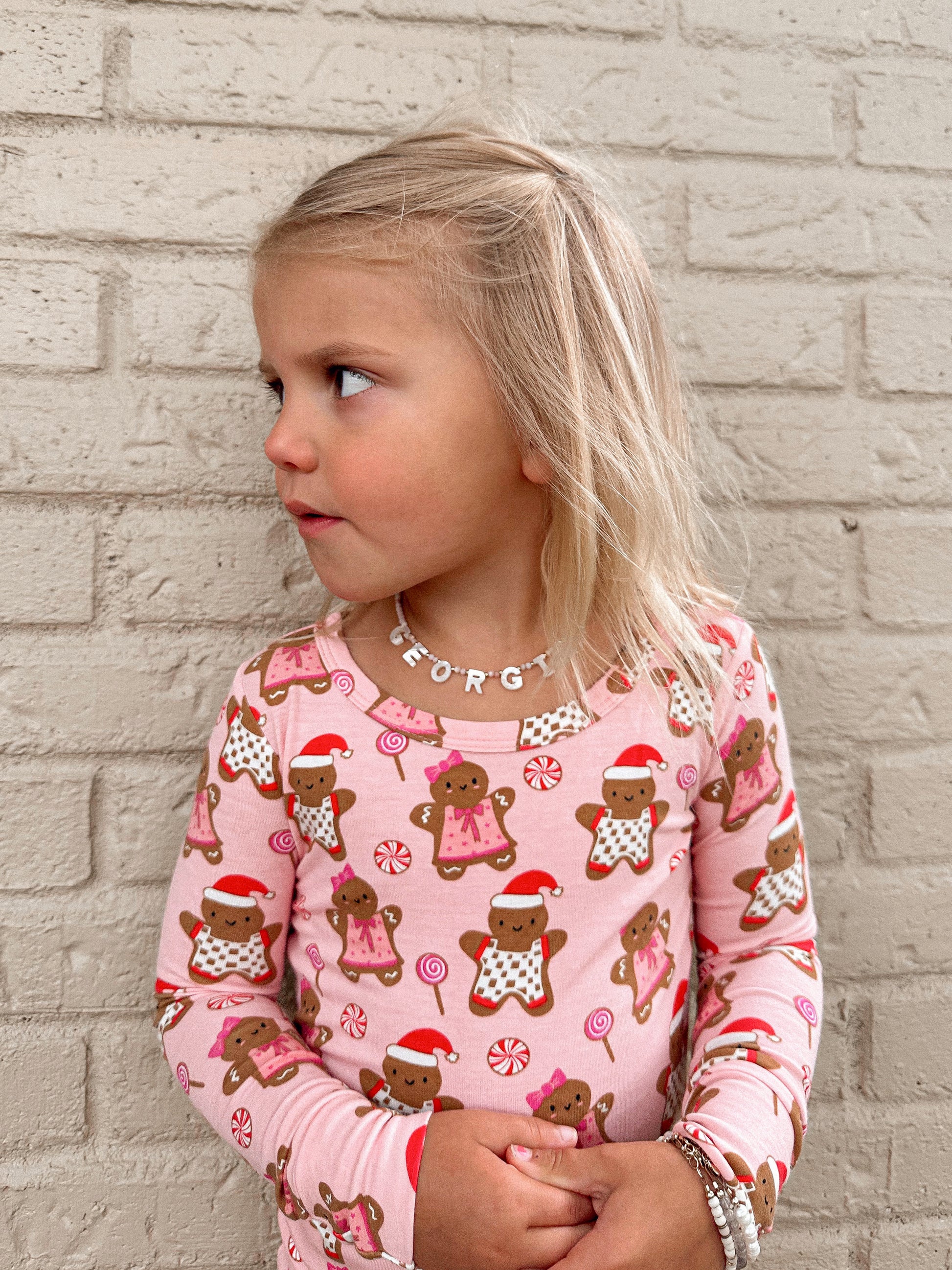 Young girl in a pink gingerbread-patterned shirt, looking sideways against a brick wall background.