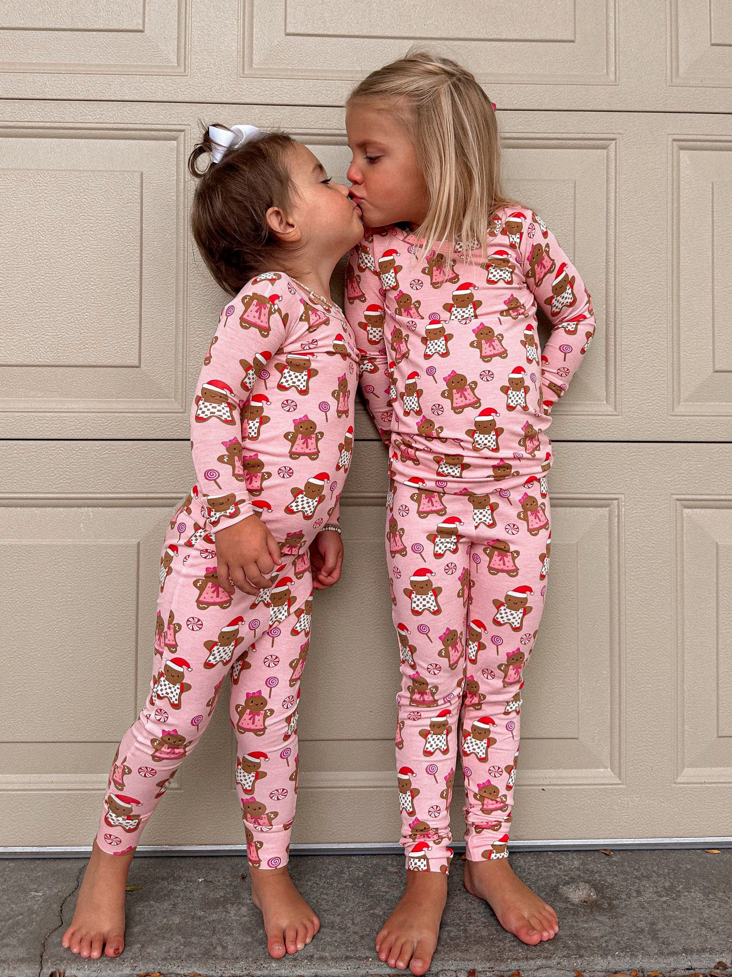 Two girls in matching pink pajamas with gingerbread patterns sharing a kiss, standing in front of a garage door.
