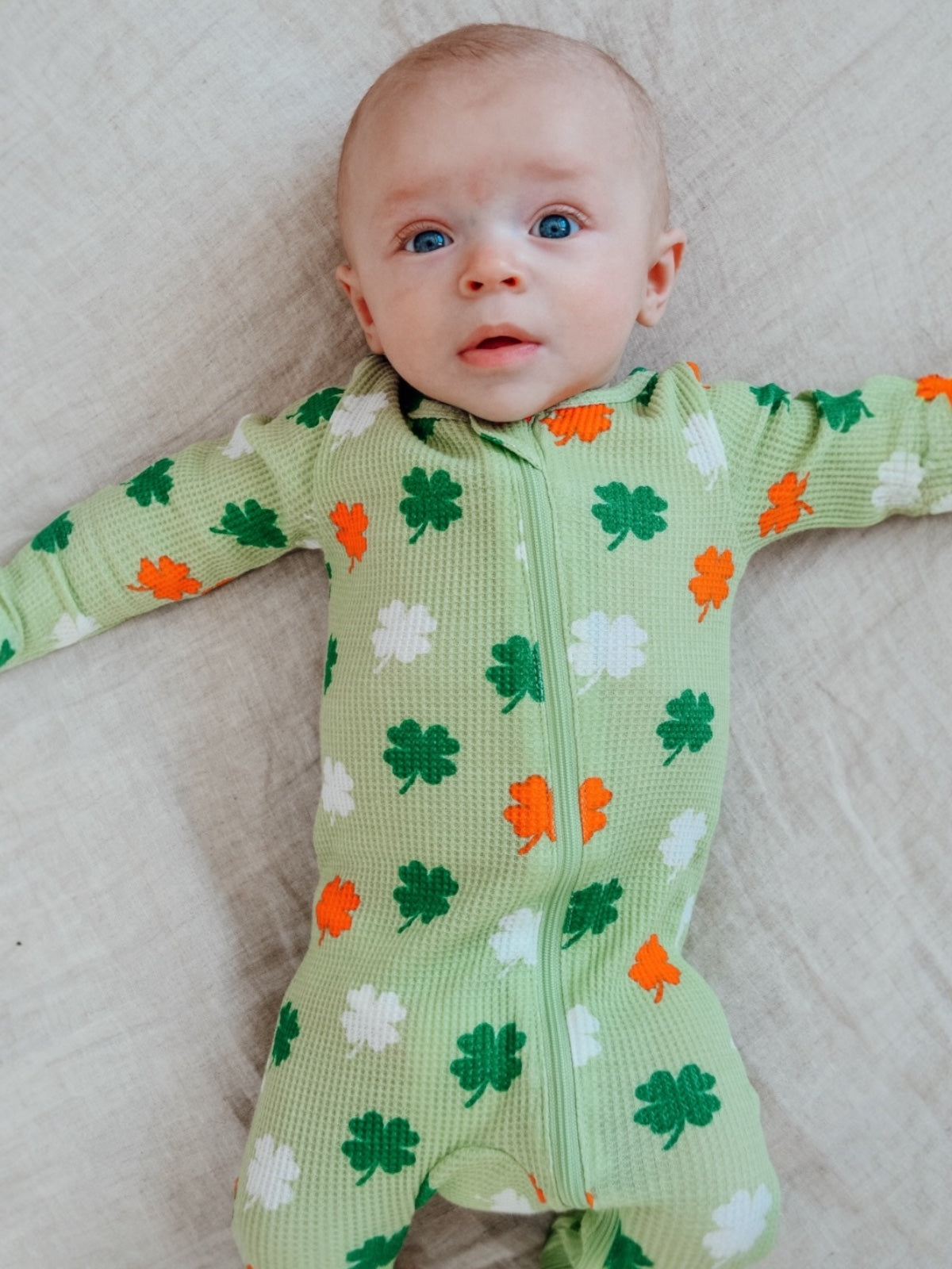Baby with blue eyes wearing a green outfit decorated with clovers, lying on a neutral-colored surface.