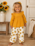 Smiling girl in a yellow top and sunflower-patterned pants stands in a cozy room with sunflowers.