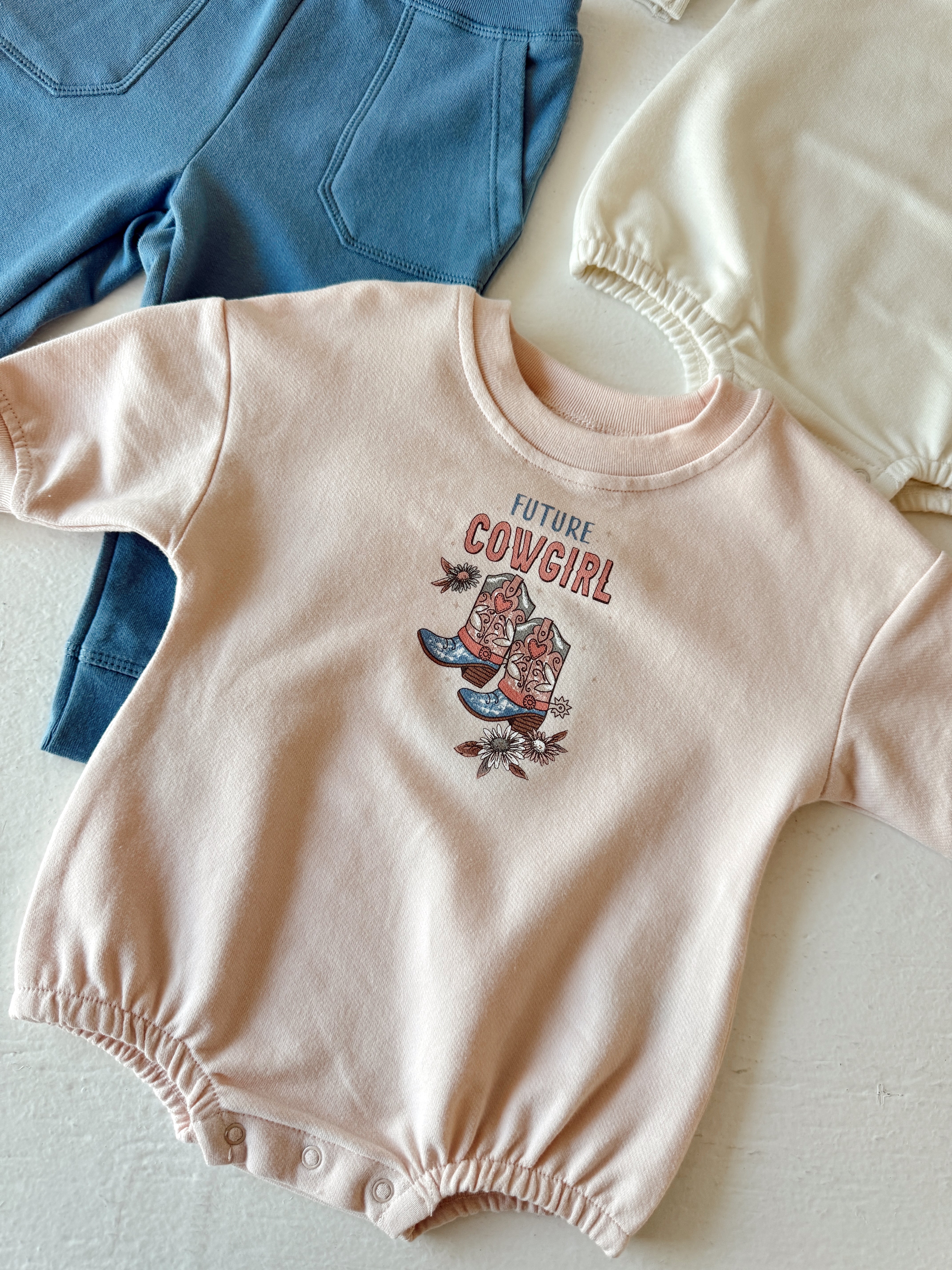 Light pink baby onesie with "Future Cowgirl" graphic, surrounded by blue and cream clothing items.