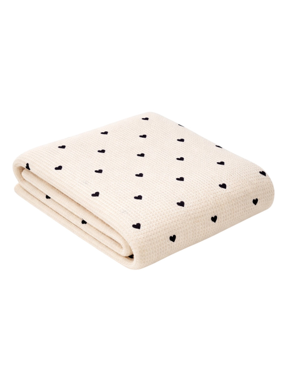 Cream-colored blanket with black heart patterns, folded neatly on a white background. Perfect for cozy settings.