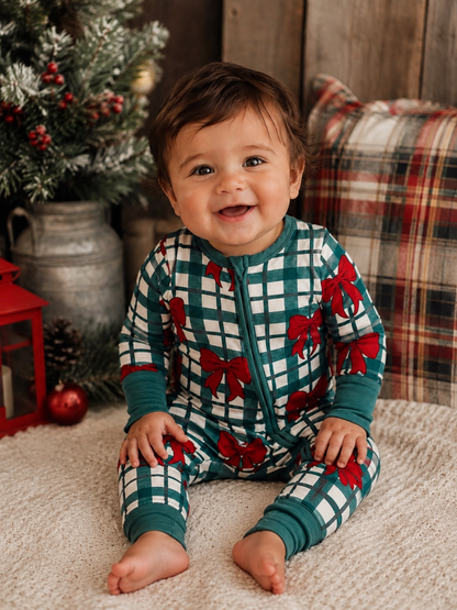 Smiling baby in festive pajamas with red bows, sitting on a cozy blanket near a decorated holiday scene.
