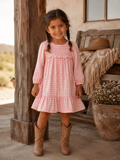 Smiling girl in a pink gingham dress and cowboy boots, standing by a rustic wooden pillar outdoors.