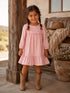 Smiling girl in a pink gingham dress and cowboy boots, standing by a rustic wooden pillar outdoors.