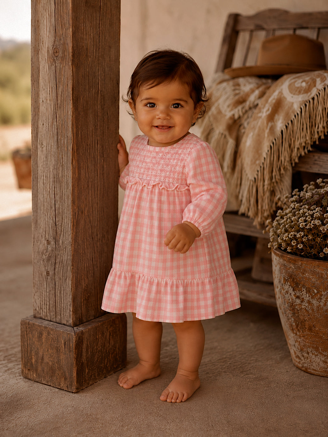 Toddler in a pink checkered dress smiles while standing by a wooden post, with plants in the background.