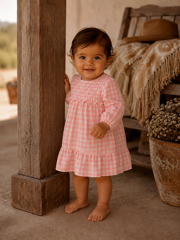 Toddler in a pink checkered dress smiles while standing by a wooden post, with plants in the background.
