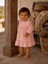 Toddler in a pink checkered dress smiles while standing by a wooden post, with plants in the background.