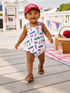 Smiling baby in a USA-themed outfit walking on a deck, with picnic items and flags in the background.