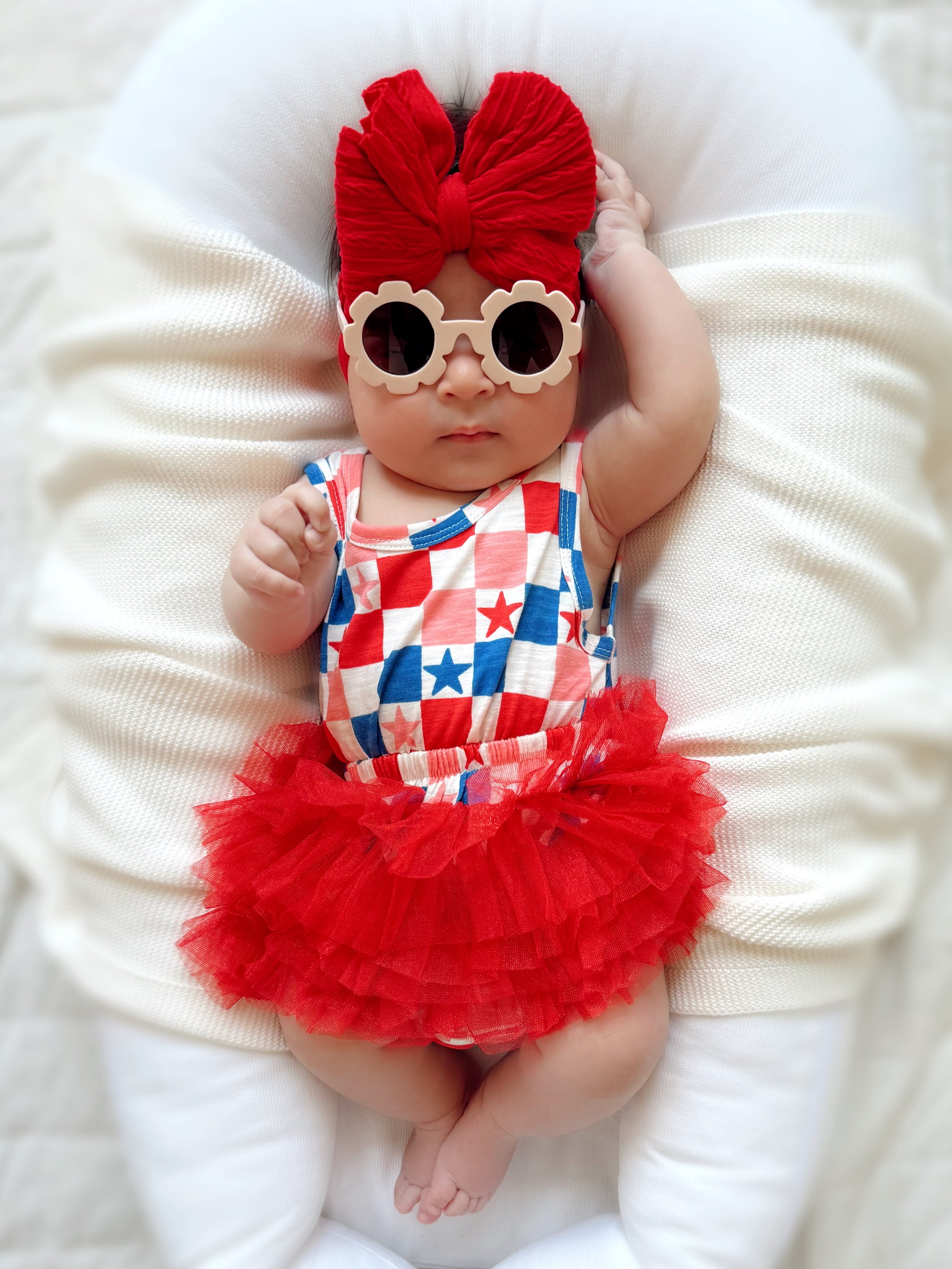 Baby in a red tutu and star-patterned outfit, wearing oversized sunglasses and a large red bow, relaxing on a soft surface.