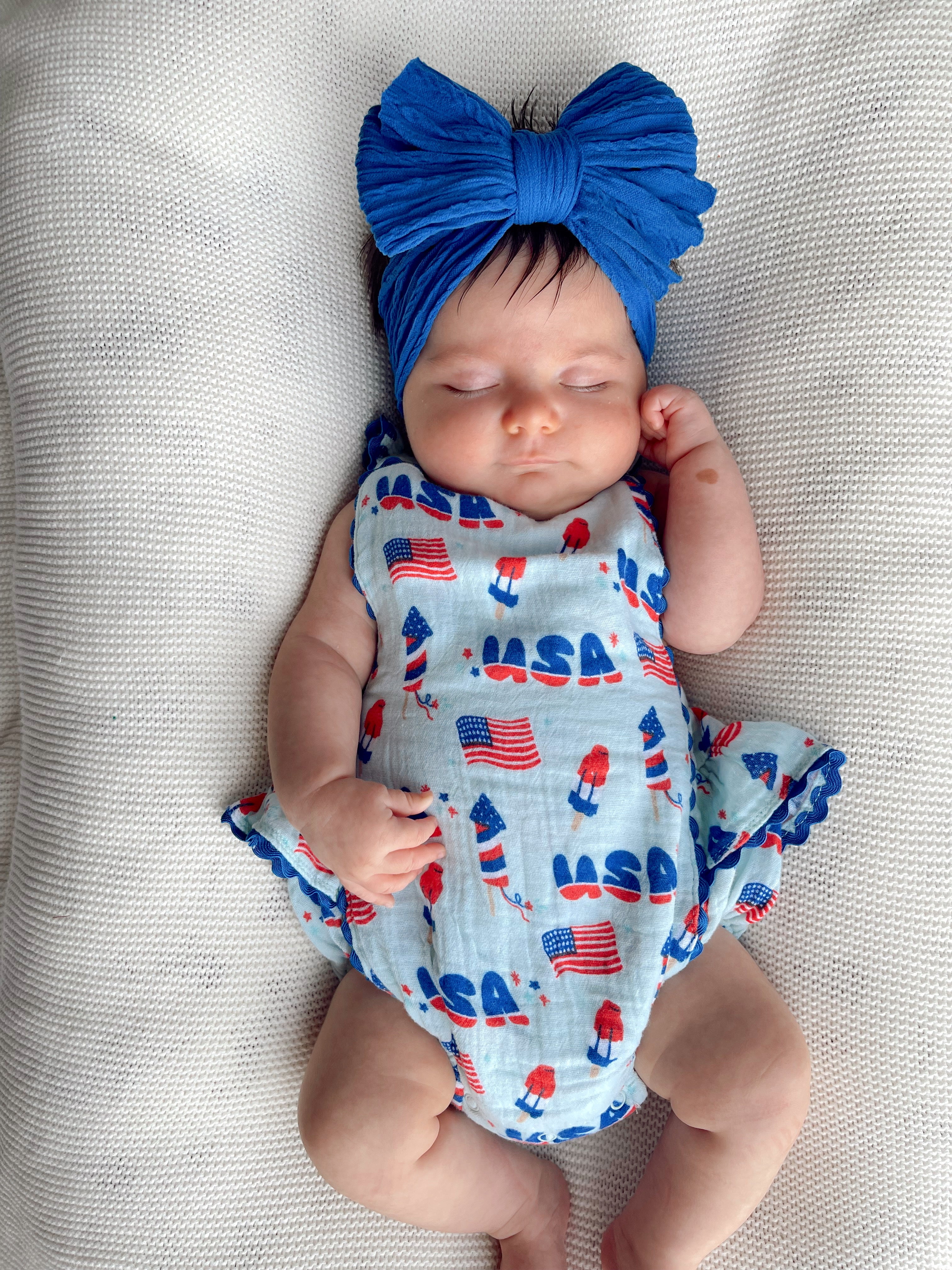 Baby sleeping on a light blanket, wearing a blue headband and a patterned onesie with rockets and American flags.