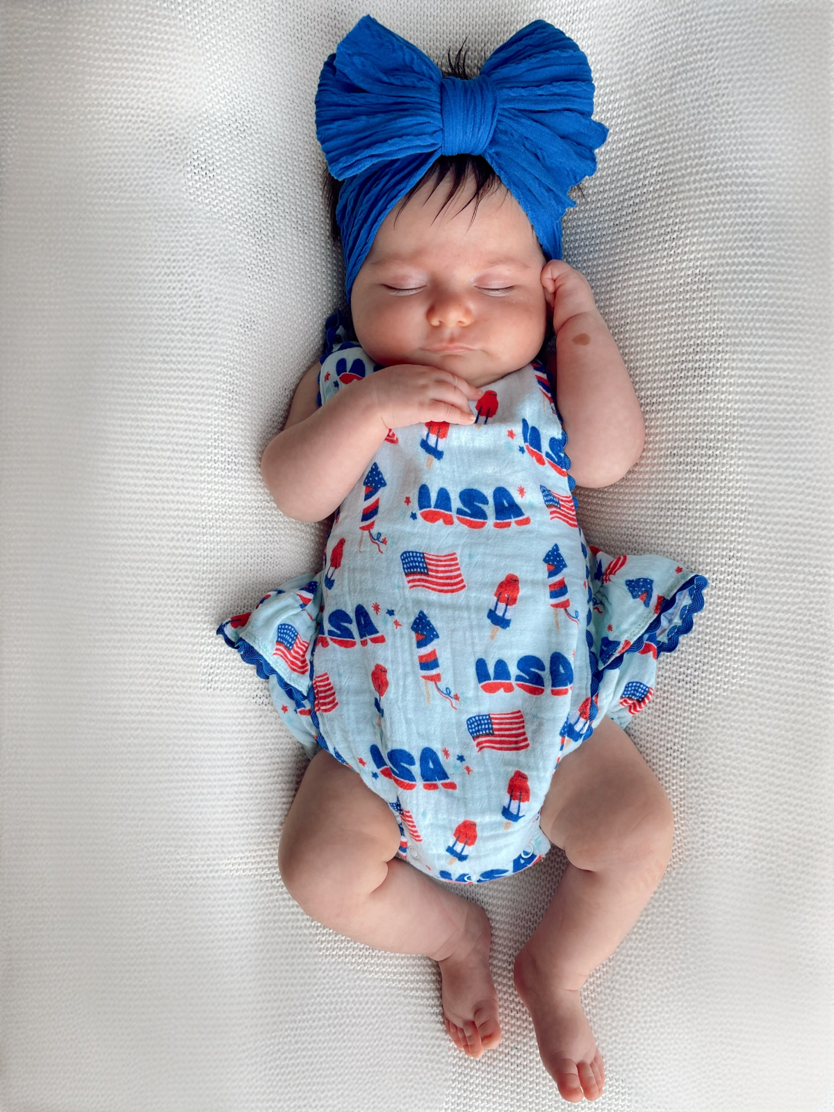 Newborn baby girl in blue headband, wearing a patriotic romper with flags and ice cream cones, sleeping peacefully.