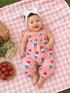 Smiling baby on a picnic blanket, wearing a pink outfit with popsicle prints, surrounded by strawberries and flowers.