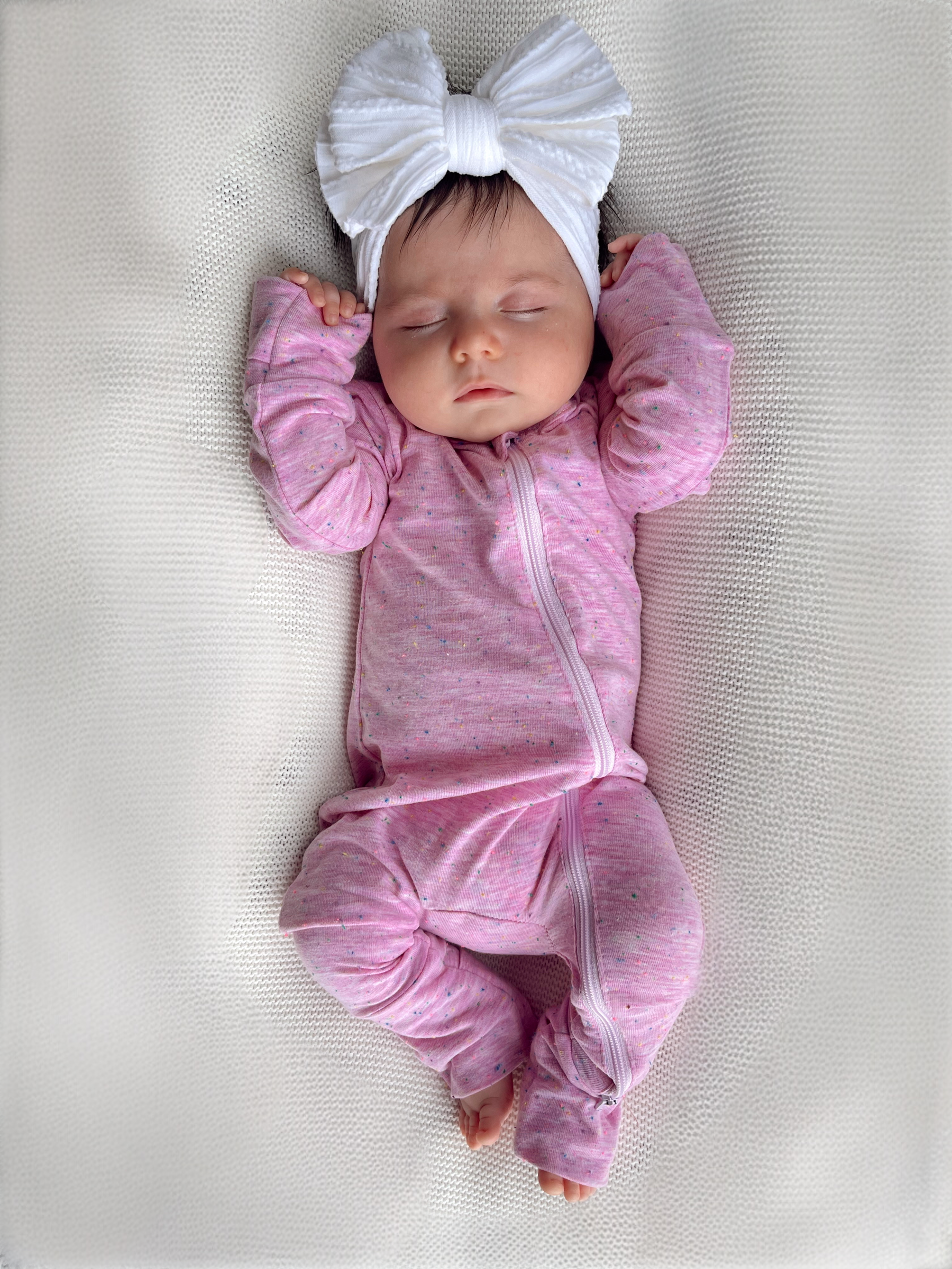 Sleeping baby girl in pink onesie with white bow headband, resting on a textured blanket.