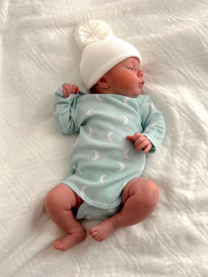 Newborn in a mint green outfit and white beanie, lying on a soft, textured blanket.