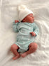 Newborn in a mint green outfit and white beanie, lying on a soft, textured blanket.