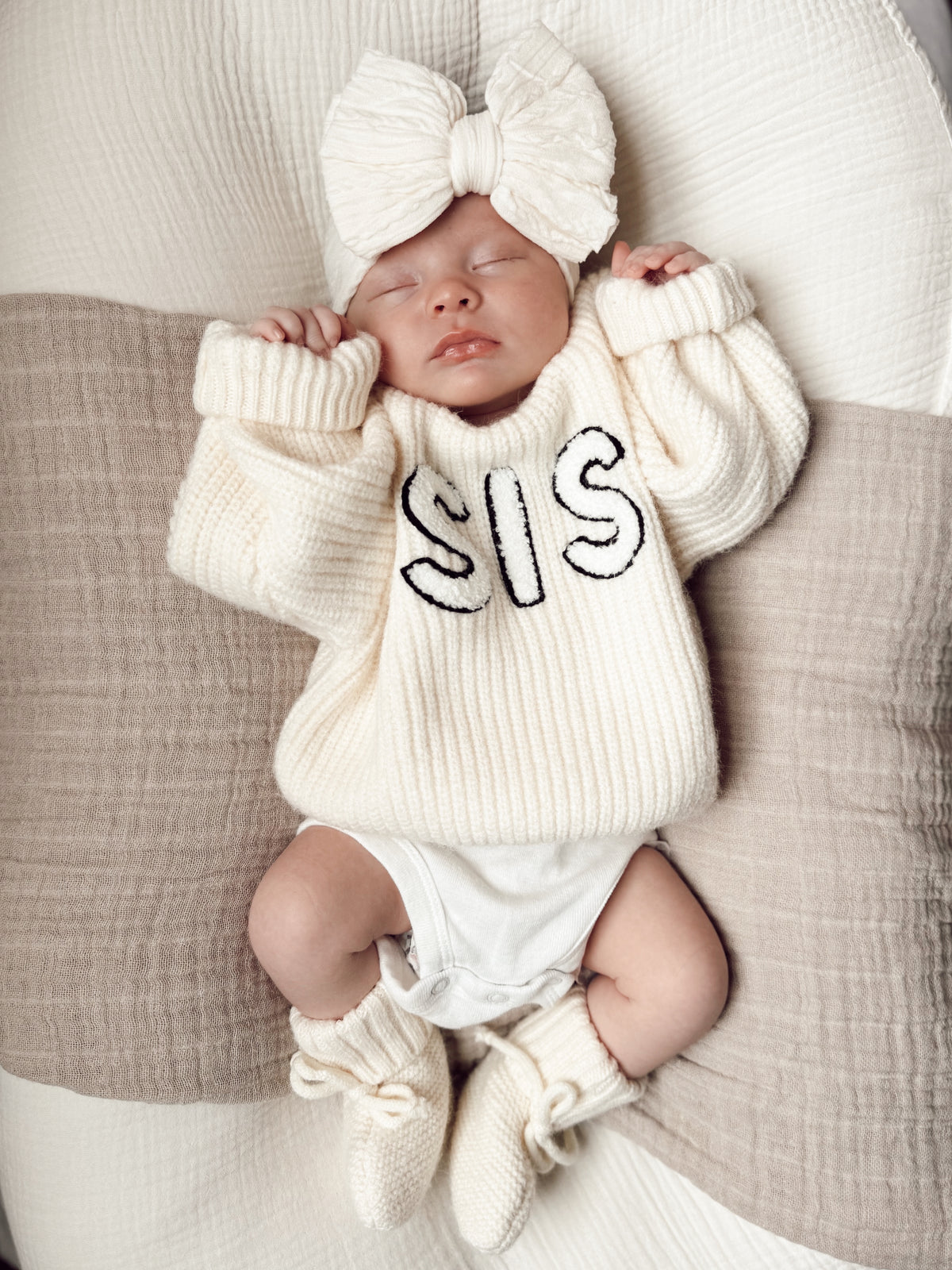 Baby girl in cozy cream sweater with "SIS" print, large bow headband, sleeping on a soft blanket.