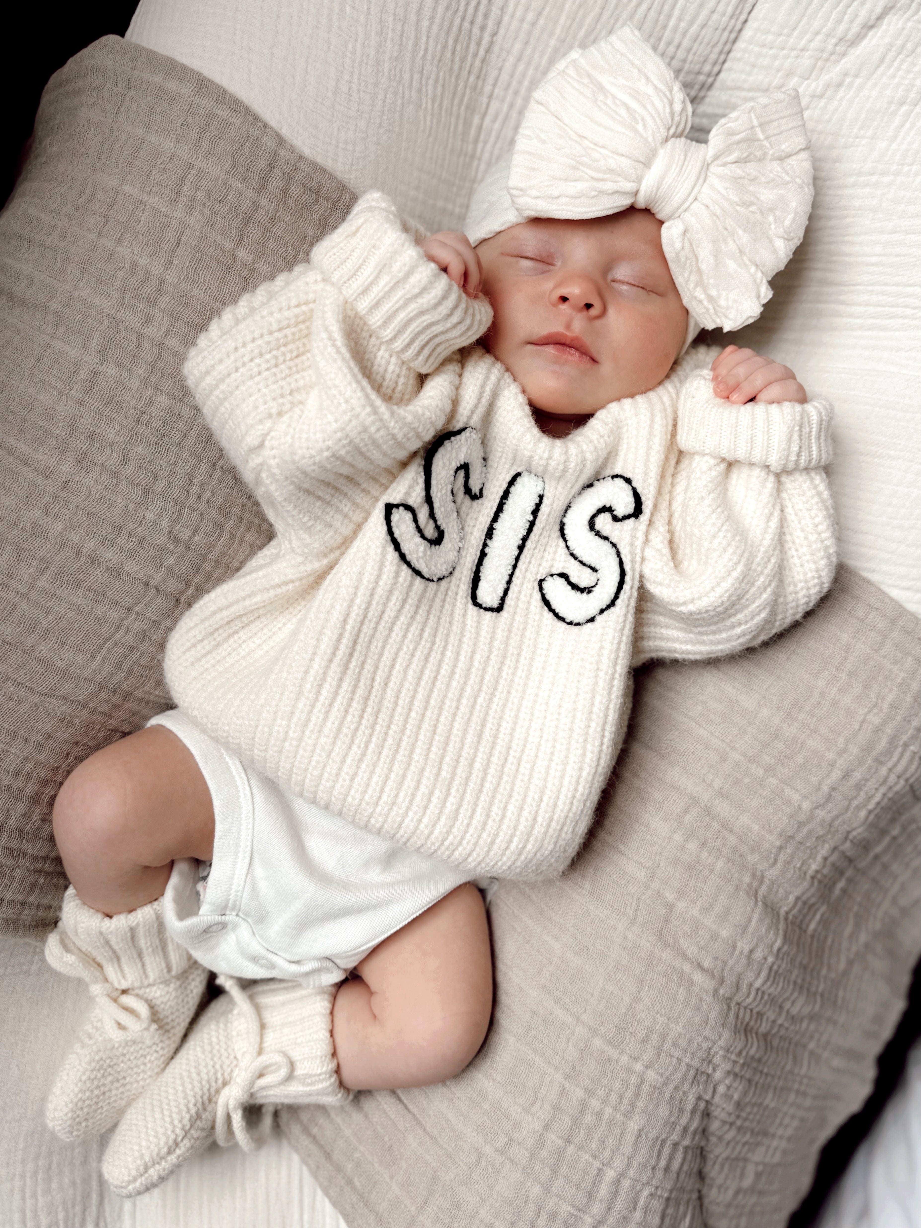 Baby wearing a cozy white sweater with "SIS" printed, resting on soft pillows with a large bow headband.