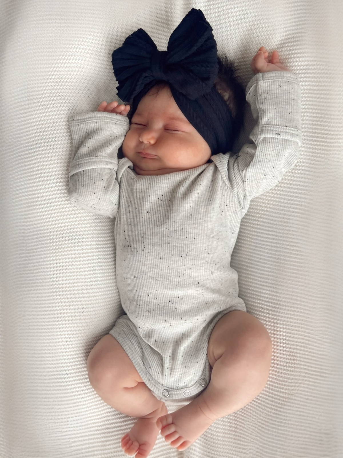 Baby girl sleeping on a textured blanket, wearing a gray onesie and a large black bow headband.