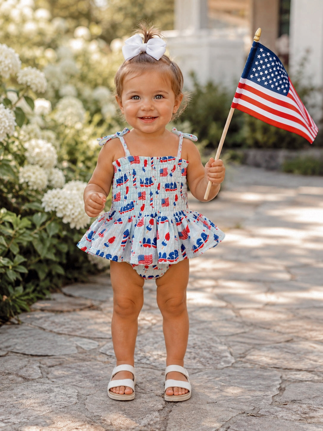Smiling child in a patriotic outfit holding an American flag, standing on a stone path surrounded by flowers.