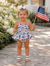 Smiling child in a patriotic outfit holding an American flag, standing on a stone path surrounded by flowers.