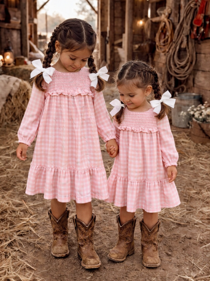 Two girls in pink gingham dresses and brown cowboy boots hold hands in a rustic barn setting.