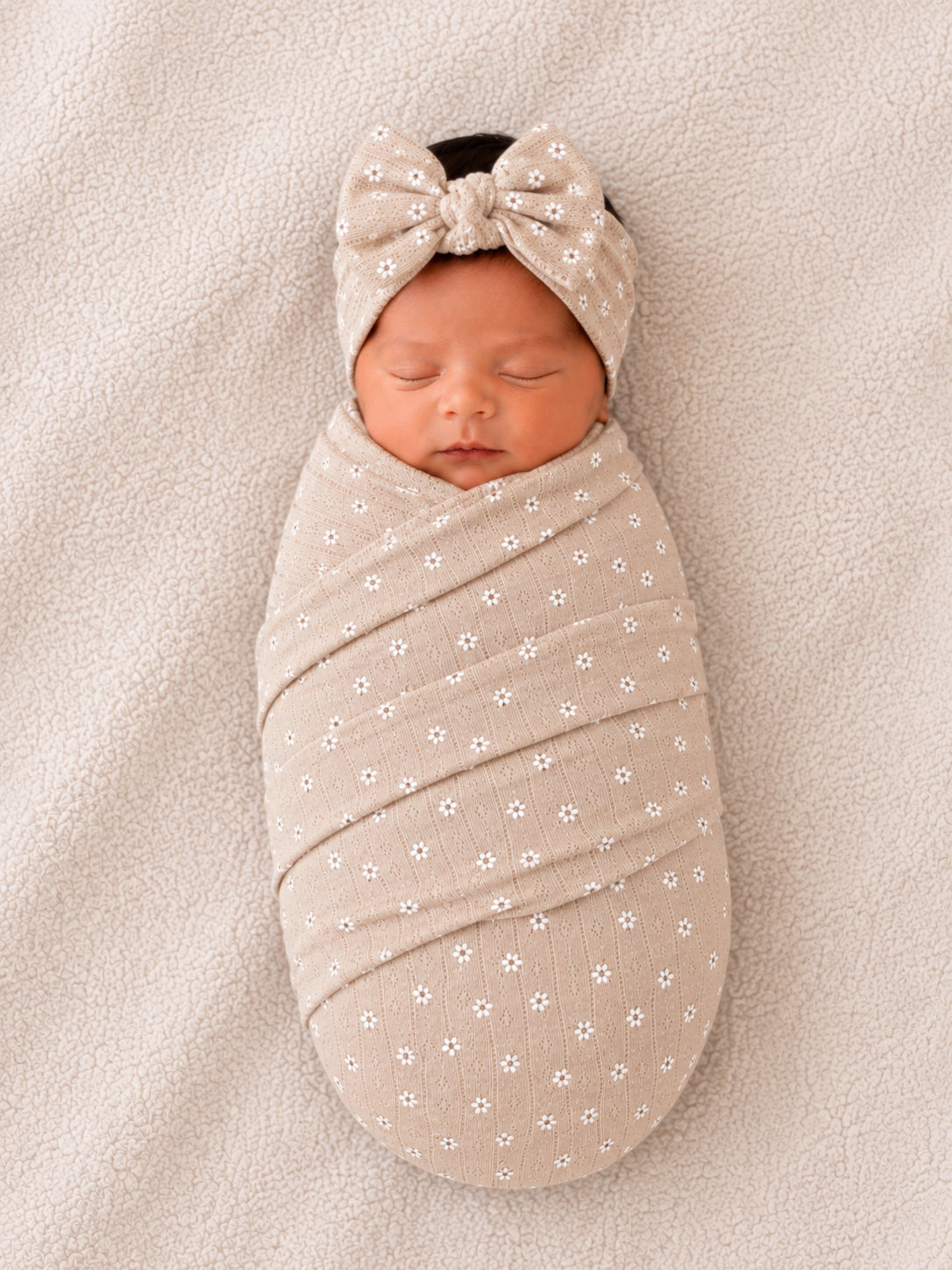 Newborn baby swaddled in a floral-patterned wrap with a bow headband, resting on soft beige fabric.
