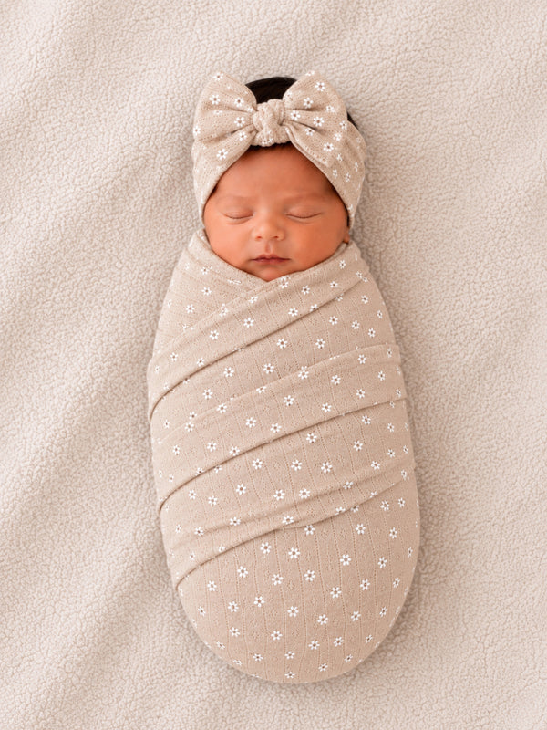 Newborn baby swaddled in a floral-patterned wrap with a bow headband, resting on soft beige fabric.