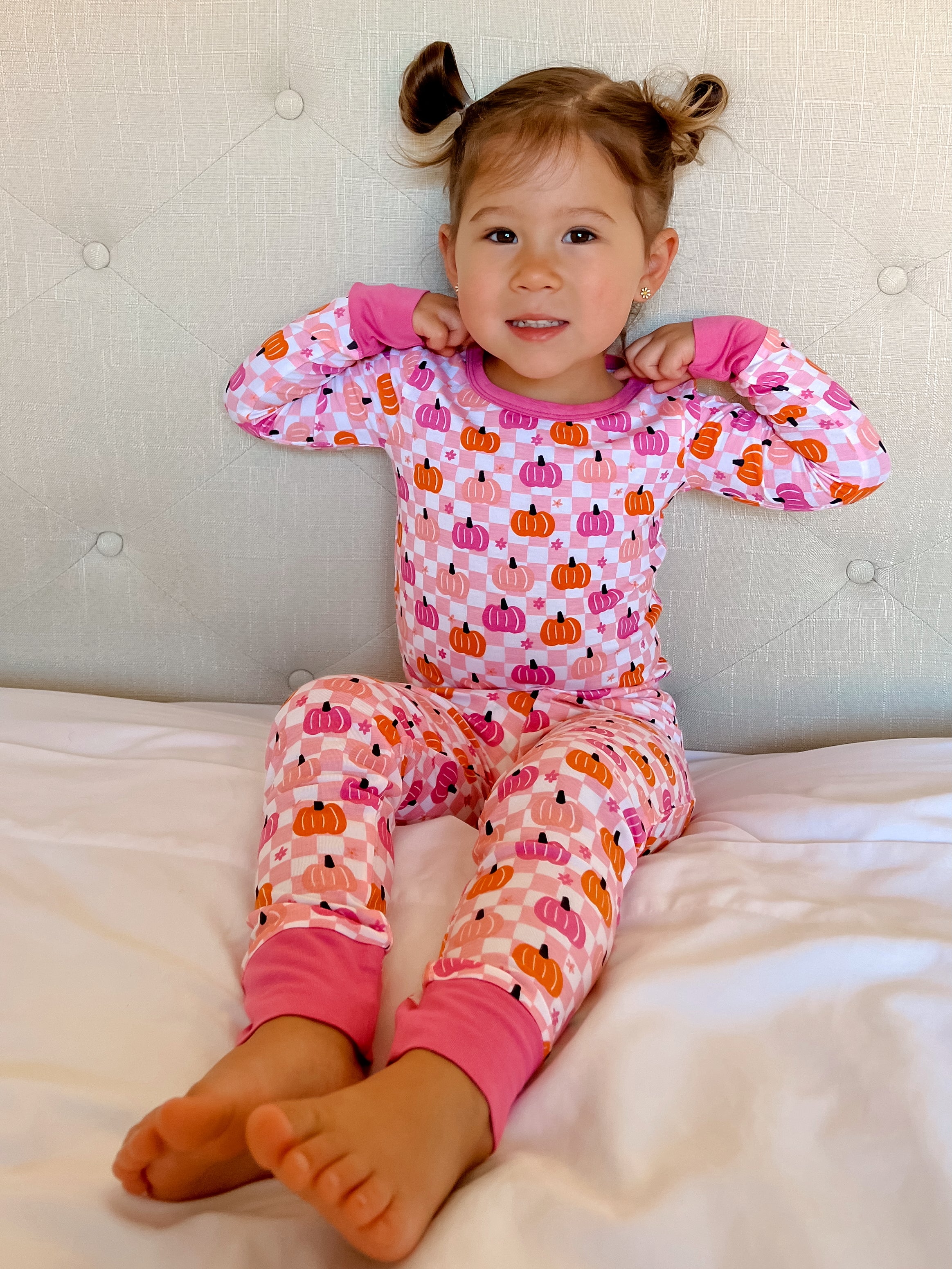Smiling child in colorful pumpkin-patterned pajamas, sitting on a bed with a light background.