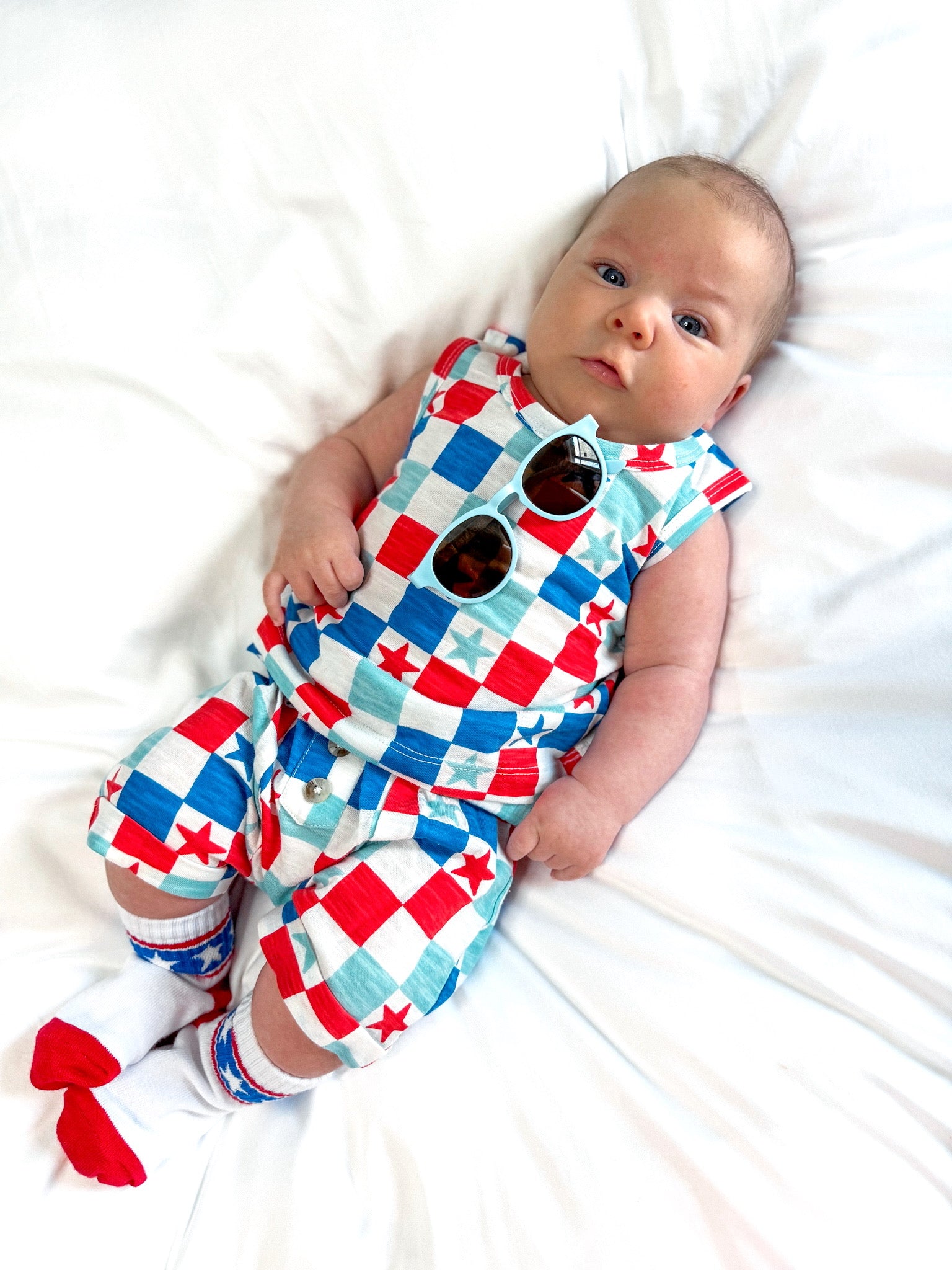 Baby wearing a colorful star-patterned outfit and sunglasses, lying on a white blanket.