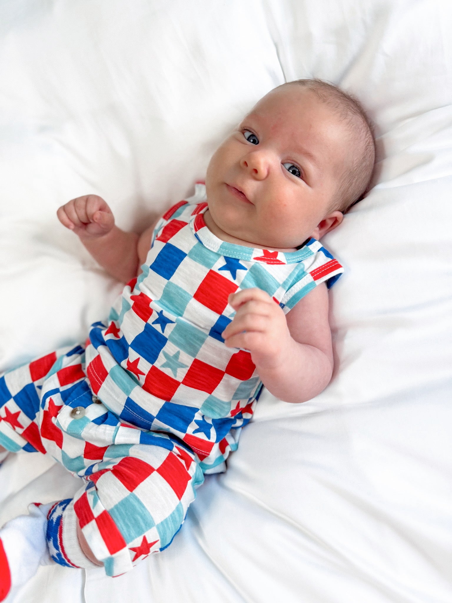 Baby in a colorful star-patterned outfit lying on a white bedspread, looking curiously at the camera.