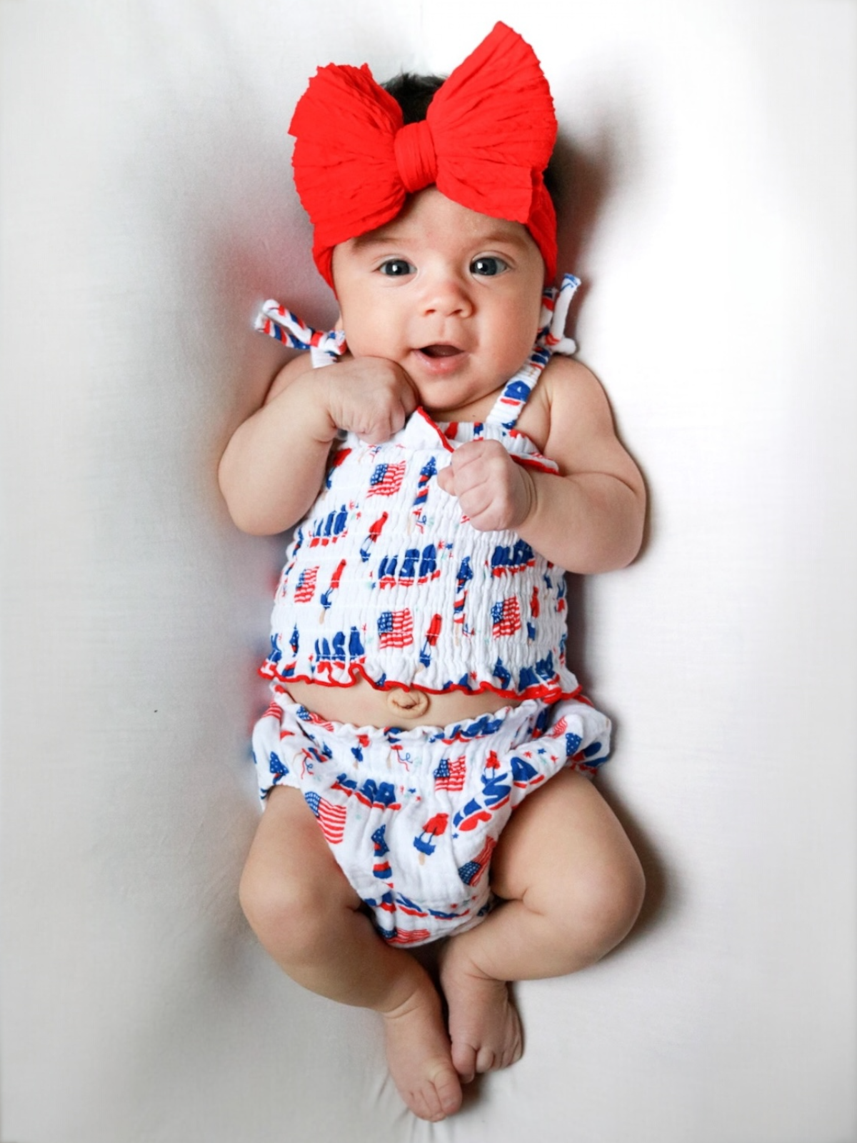 Smiling baby dressed in a patriotic red, white, and blue outfit with a large red bow on their head.