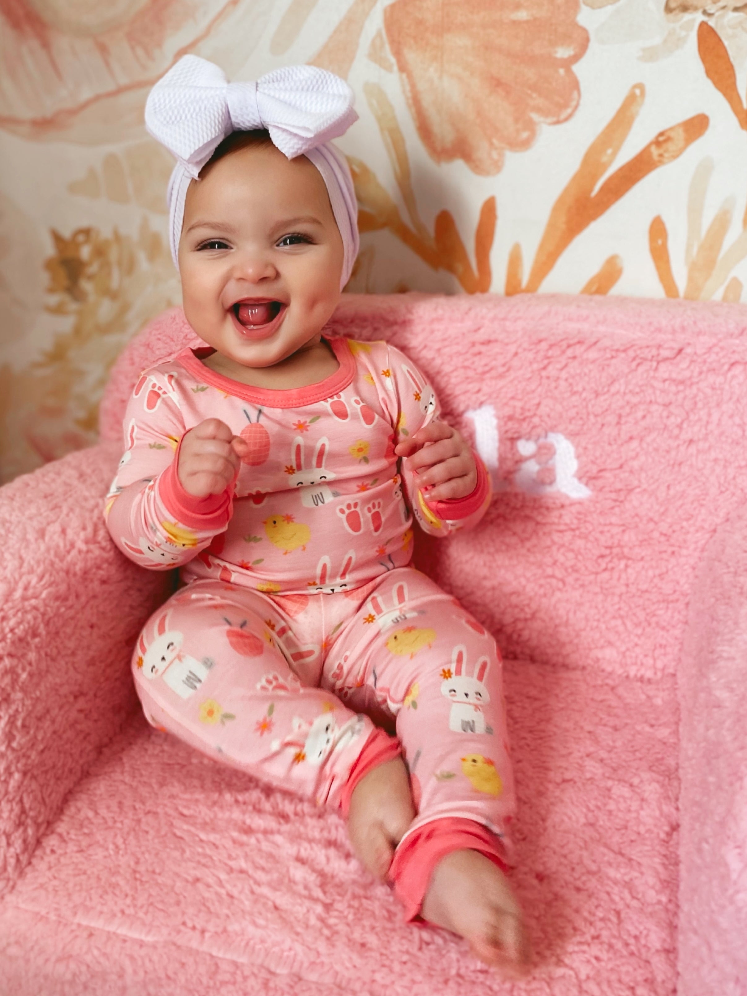 Smiling baby in colorful pajamas with a pink headband, sitting on a plush pink chair against a floral background.