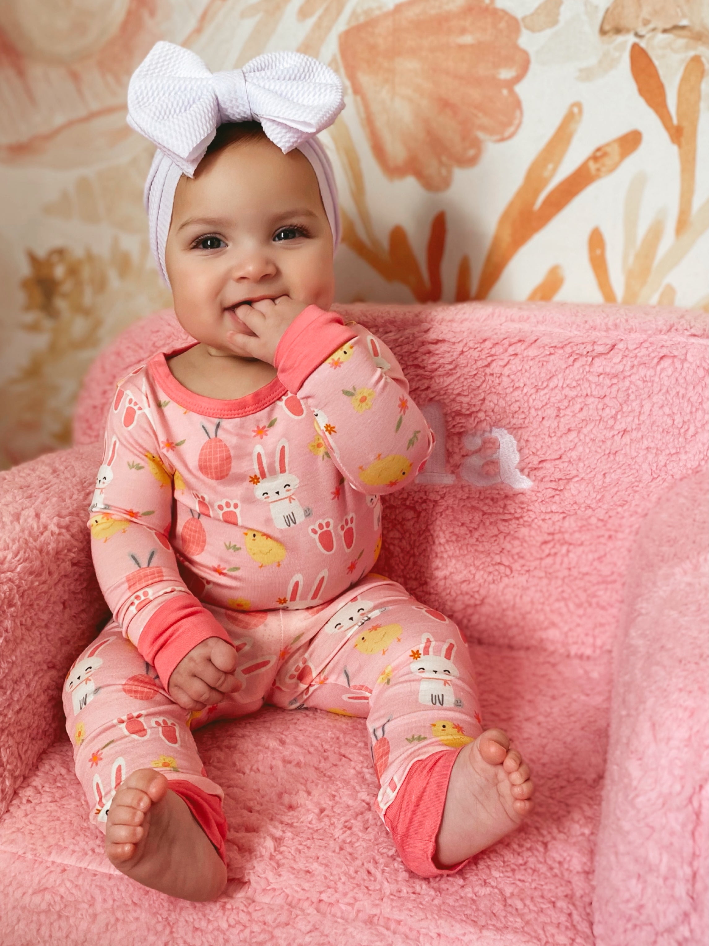 Smiling baby in pink patterned pajamas sits on a soft pink chair, playing with her hand and wearing a bow headband.