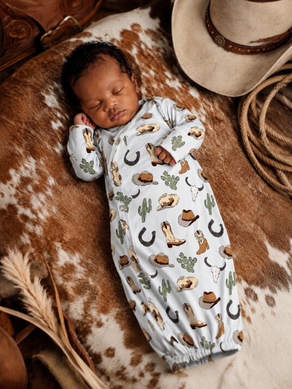 Sleeping baby in cowboy-themed onesie, nestled on a patterned blanket with a hat and cowboy accessories nearby.
