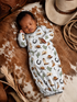 Sleeping baby in cowboy-themed onesie, nestled on a patterned blanket with a hat and cowboy accessories nearby.