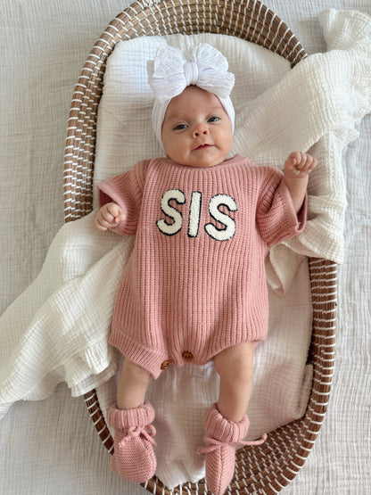 Baby in pink knit romper with "SIS" text, wearing a large white bow and booties, lying in a woven basket.