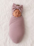 Newborn baby swaddled in a soft pink ribbed wrap, wearing a matching oversized bow headband, sleeping peacefully on white fabric.