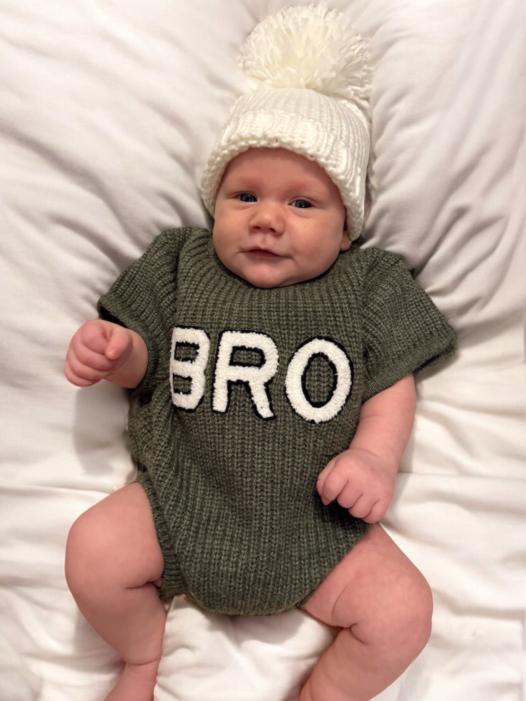 Baby wearing a green sweater with "BRO" printed on it, posing on a white blanket, topped with a white pom-pom hat.