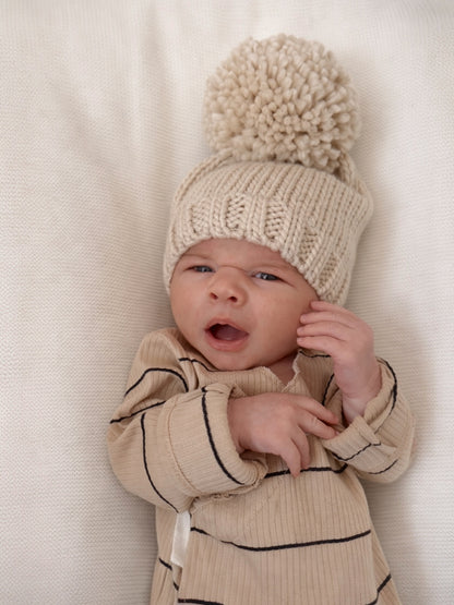 Baby wearing a beige knitted hat with a pom-pom, yawning on a light-colored blanket.