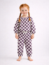 Smiling child with braided hair wearing a purple and white checkered outfit, standing barefoot on a light background.