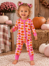 Smiling toddler in pumpkin-patterned pajamas stands on a cozy rug with pumpkins and flowers in the background.