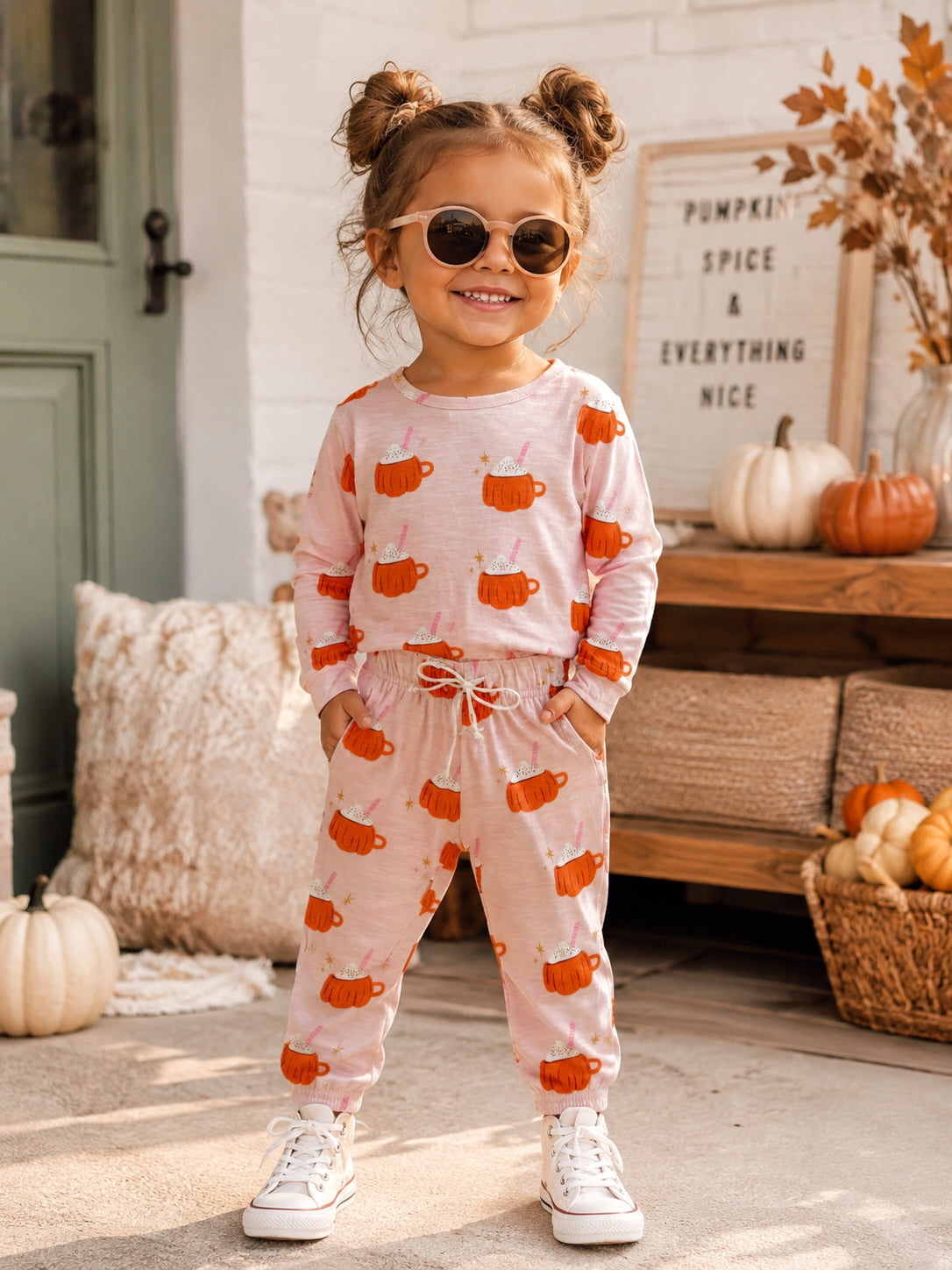 Smiling child in pumpkin-themed outfit, wearing sunglasses, standing in cozy fall setting with pumpkins.