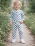 Smiling child in blue and white checkered outfit, standing on a path surrounded by greenery and wildflowers.