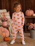 Smiling girl in cozy pink pumpkin-patterned outfit, standing among pumpkins and flowers, in a warm, autumn setting.